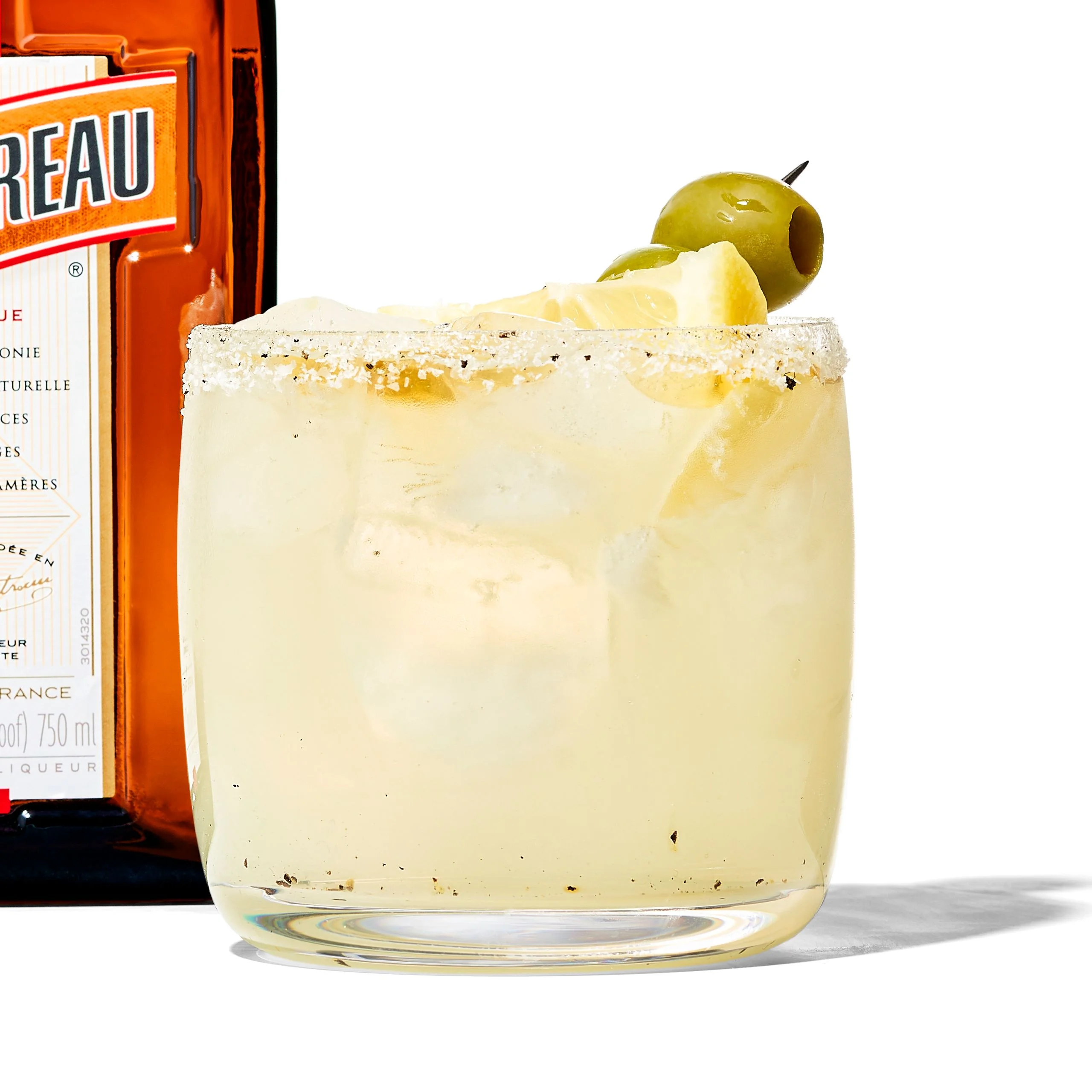 A mexcal margarita with Cointreau in a rocks glass garnished with black pepper and an olive and lemon wedge skewer