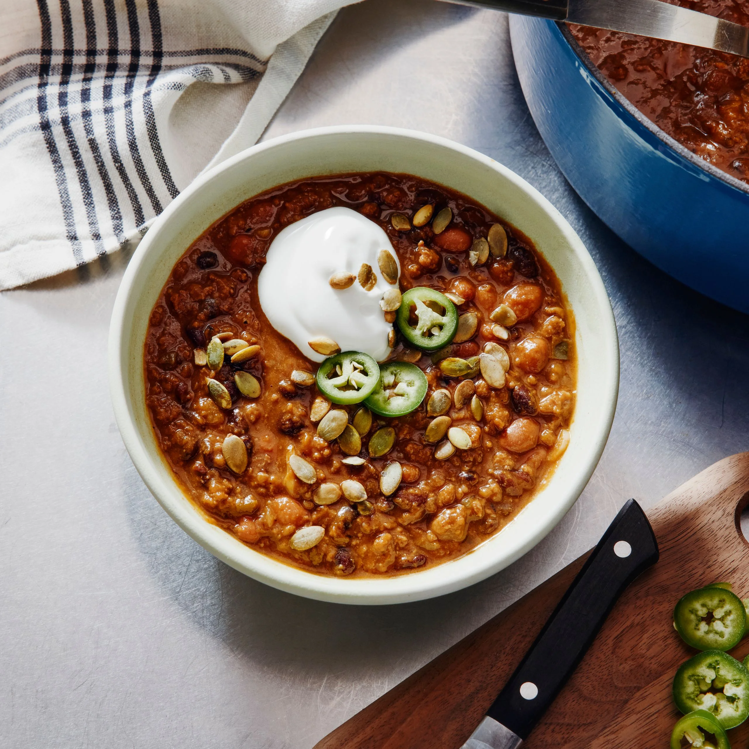A single serving of spiced beef and pumpkin chili alongside the full yield and a cutting board with jalapeño slices.